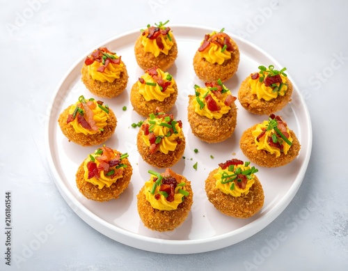 Deviled eggs, breaded and fried, topped with bacon and chives, arranged on a white plate