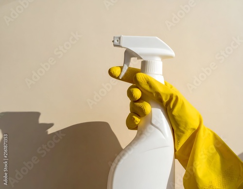 Close-up of a hand in yellow glove holding a white spray bottle against a light wall