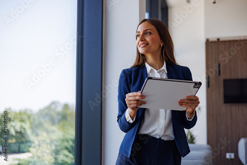 Fotografie Businesswoman holding tablet, smiling and looking through office window, envisio
