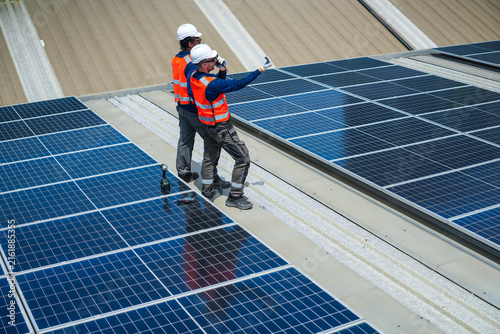 Canvas Print Two rooftop solar energy workers in protective safety gear methodically stride across the finished rows pointing out final checks on a large corrugated factory rooftop under the tropical