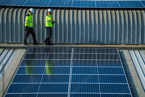 A bearded engineer and his colleague in reflective vests and helmets calmly walk the roof walkway beside the darkening array on the sweeping metal roof of a manufacturing plant in Southeast.