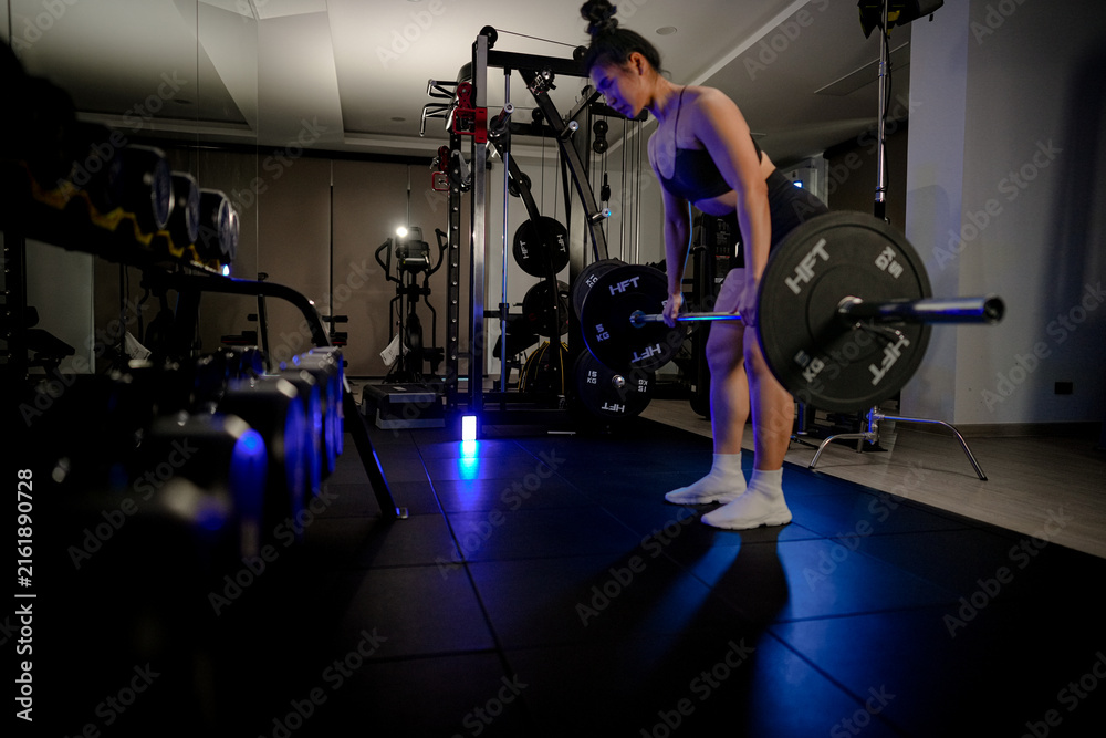 Fototapeta premium Empowered women in the gym: a determined Asian woman setting up for a strong barbell deadlift in a neon-lit weight room, lit by dramatic low-key lighting, with unwavering focus