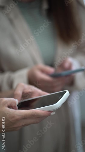 Closeup of woman manicured hands holding white smartphone, another person using phone in soft-focus background. Modern business lifestyle and technology.