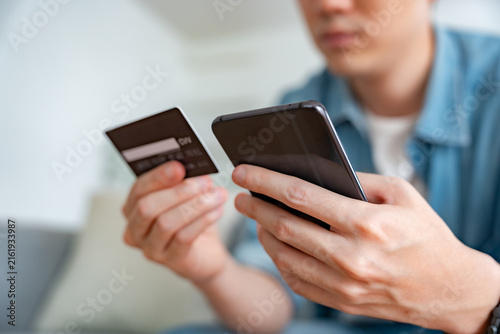 Close-up image of a man's hand holding a credit card and using a smartphone to make online purchases via internet banking in his living room at home