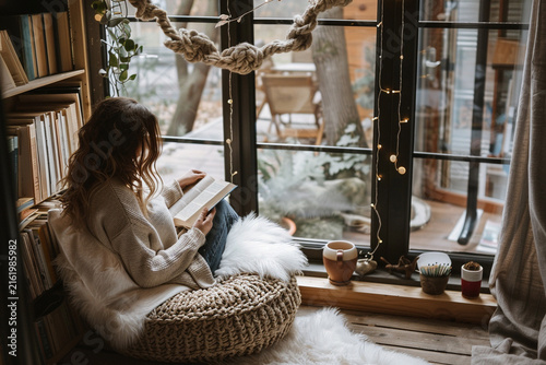 Young woman reading a book in a cozy corner on a fluffy pouf seat near a window with fairy lights and bookshelves