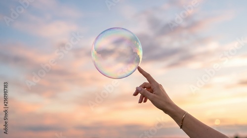 Hand gently touching iridescent soap bubble against soft pastel sunset sky, delicate moment of fragility and beauty, childhood wonder, dream, ephemeral nature of life and fleeting
