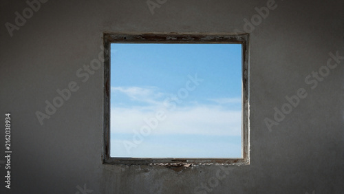 Simple square window on a weathered concrete wall framing a clear blue sky with white clouds