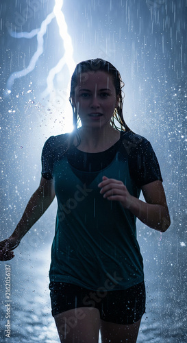 Young athletic woman running through heavy rain shower with bright lightning bolt striking in background night sky during dramatic storm