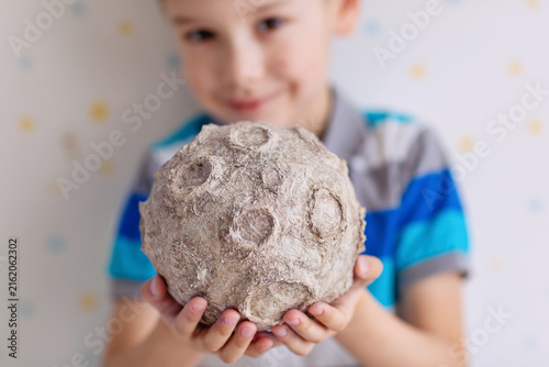 A cheerful young boy in a striped polo gently cradles a textured papier‑mâché moon with realistic craters. Soft studio lighting highlights the tactile surface against a subtly patterned backdrop.