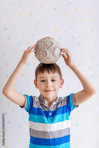 A cheerful young boy in a striped polo balances a textured papier‑mâché moon atop his head. The playful scene features a white backdrop dotted with pastel stars and soft studio lighting.