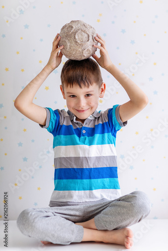 A cheerful young boy in a striped polo shirt sits cross‑legged balancing a textured papier‑mâché moon atop his head. The playful scene features a star‑patterned backdrop and soft studio lighting.