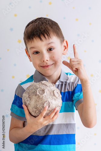 A cheerful young boy in a striped polo holds a textured papier‑mâché moon. He smiles and points upward against a white backdrop dotted with yellow and blue stars, conveying playful curiosity.