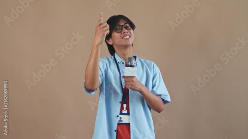 Young Asian male reporter with glasses holding microphone and counting to three with fingers in studio