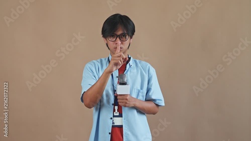 Young Asian male reporter holding microphone and making quiet shush gesture with finger to lips in studio