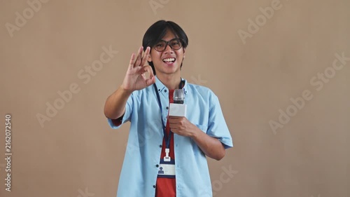 Young Asian male reporter holding microphone and smiling while making OK hand gesture in studio setting