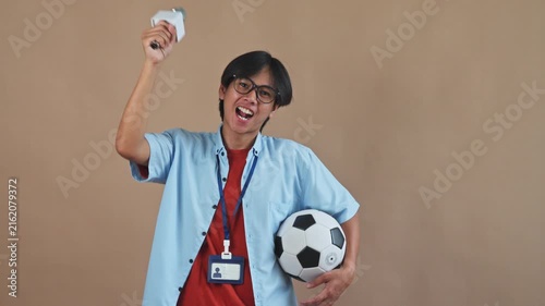 Excited Asian sports reporter holding soccer ball while raising microphone and cheering passionately in studio