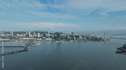 Aerial view of Ha Long waterfront in Quang Ninh, Vietnam, showing the calm bay, marina, city skyline, coastal architecture, and surrounding mountains under a blue sky.