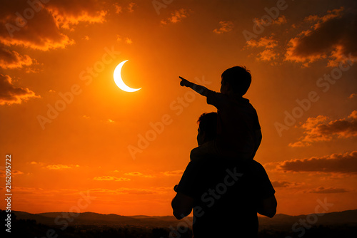 Silhouette of child sitting on parent shoulders pointing at solar eclipse in golden sunset sky
