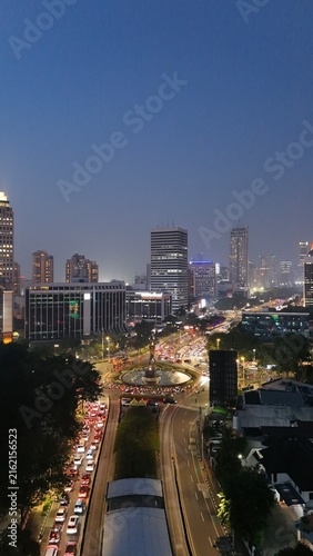 Aerial drone view of Jakarta's Sudirman business district at night featuring illuminated skyscrapers, glowing office buildings, vibrant city lights, and heavy traffic after office hours.