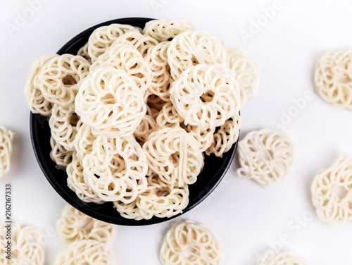 Overhead view of a black bowl filled with raw white kerupuk udang (Indonesian prawn crackers) with several loose pieces scattered on a clean white background, showcasing a textured food composition.
