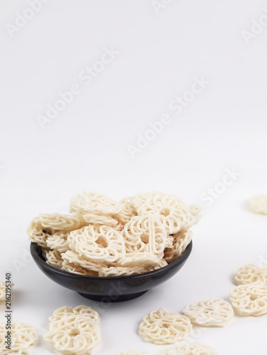 Front-angle view of a black bowl filled with raw white kerupuk udang (Indonesian prawn crackers) with loose crackers scattered on a clean white background