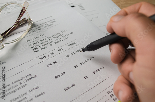 Close-up of a person completing an invoice with a black pen, surrounded by paperwork and eyeglasses. Business accounting, billing, finance, payment and office work concept.