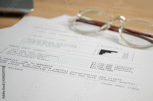 Business financial report document with numbers and charts lying on a wooden office desk next to reading glasses, representing investment analysis, corporate accounting, and audit concepts. Copy space