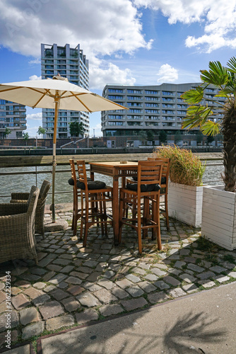 Cozy outdoor cafe terrace with wooden table and umbrella by the river. Summer waterfront restaurant patio with modern city architecture skyline on background. Beautiful sunny day vacation concept.