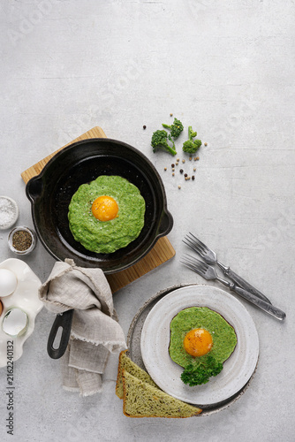 Healthy green fried egg with broccoli puree yolk inside in a cast iron skillet. Top view of a nutritious breakfast flat lay with whole grain bread on concrete background.