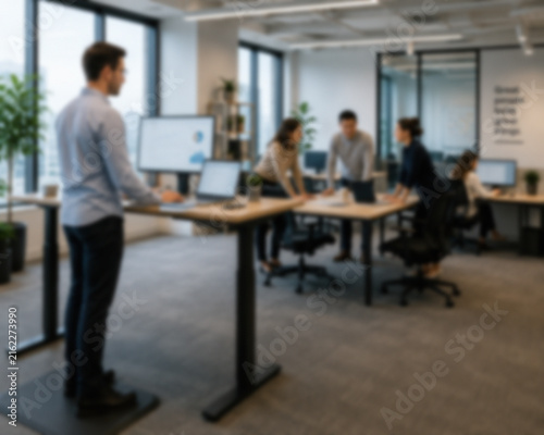 Blurred Employee Using A Standing Desk While Coworkers Collaborate In A Modern Office
