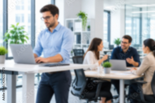 Blurred Employee Working At A Standing Desk While Coworkers Collaborate In A Modern Workplace