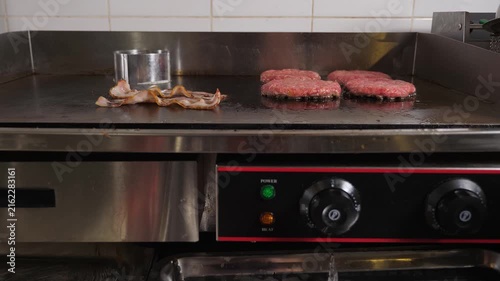 Close-up of a chef frying bacon and hamburger patties on a large stove in a restaurant kitchen. The chef is cooking a juicy cutlet and bacon to make a delicious burger.