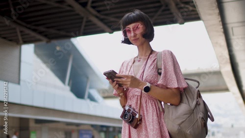 A female traveler with a camera and a backpack stands on a railway station platform, smiling. She is looking at her smartphone and waiting for the train; the train schedule is on the app.
