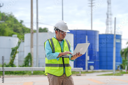 Industrial engineer hard hat safety vest laptop water tank utility plant worker inspection maintenance outdoor male technician at blue facility site