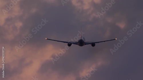 Airplane flying away into cloudy sunset sky with wingtip navigation lights glowing during twilight, rear view tracking shot.