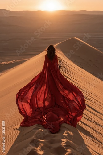 Woman in red dress standing on sand dune at sunset in desert