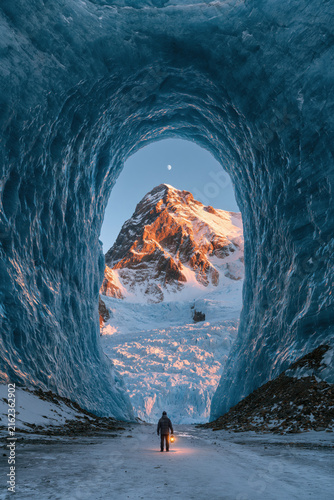 A person walks through an ice tunnel with large blue walls and a mountain in the background during sunset