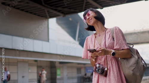 A female traveler with a camera and a backpack stands on a railway station platform. She is looking at her smartphone and waiting for the train. Checking the train schedule in an app. 

