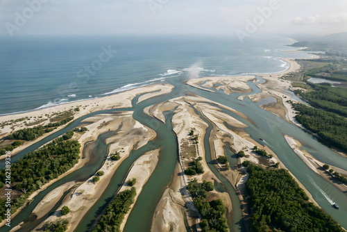 Angle drone view of a river delta flowing into the sea with intricate sand patterns, green mangroves, and blue ocean waves at the coast