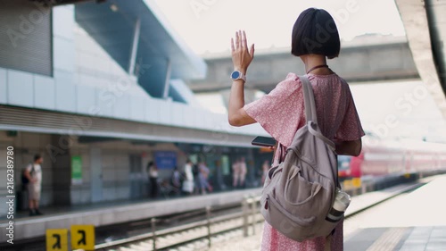 The girl in a pink dress and with a camera stands on the platform of the train station and waves with her hand. goodbye to friends. travel and vacation. creating content about travel. tourist with cam