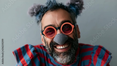 Laughing bearded clown portrait with gray pom pom hair and red striped costume wearing round red glasses for expressive editorial portrait background