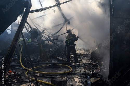 Firefighter in protective gear extinguishes a structural fire with a high-pressure fire hose amid dense smoke, flames, twisted metal debris, collapsed structures, and hazardous conditions.