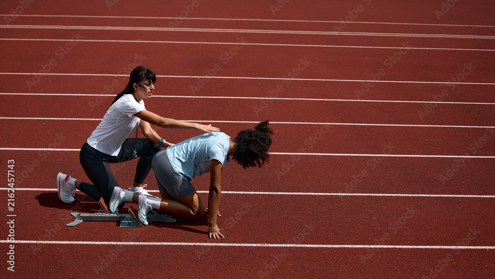 Fototapeta premium Adult female coach correcting teenage runner crouched starting position on track. Concept of hands on training assistance, athlete safety instruction, summer sport mentorship.