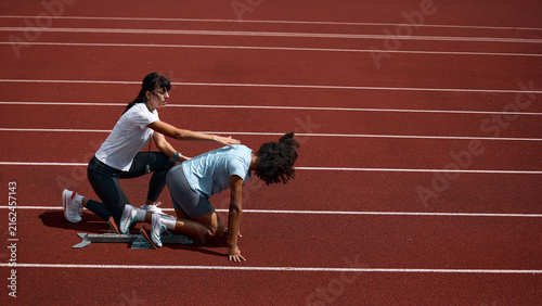 Adult female coach correcting teenage runner crouched starting position on track. Concept of hands on training assistance, athlete safety instruction, summer sport mentorship.