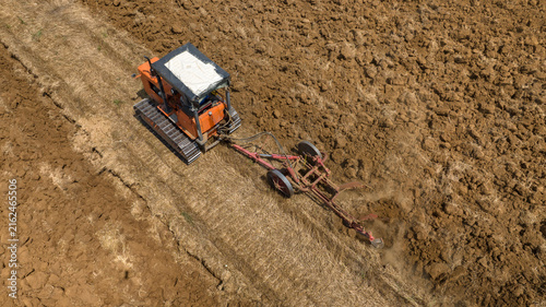 Photography Aerial closeup of a orange tracked bulldozer moves across a dry dirt field, towing a long metal agricultural attachment behind it