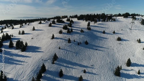 Aerial view of Kopaonik mountain slopes covered in snow with scattered pine trees and ski lift infrastructure under a clear blue sky in Rasina District, Serbia.