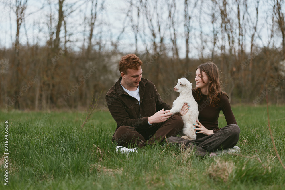 Fototapeta premium Casual outdoor lifestyle with a cheerful couple sitting in a field, playing with a goat kid, warm afternoon light, relaxed smiles, gentle interaction, natural setting, candid moment of joy and