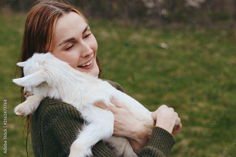 Fototapeta premium Lifestyle woman goat kid outdoors affection cuddle nature happiness photography. A candid scene of a smiling woman holding a goat kid in a grassy field, celebrating connection and gentle bonding.