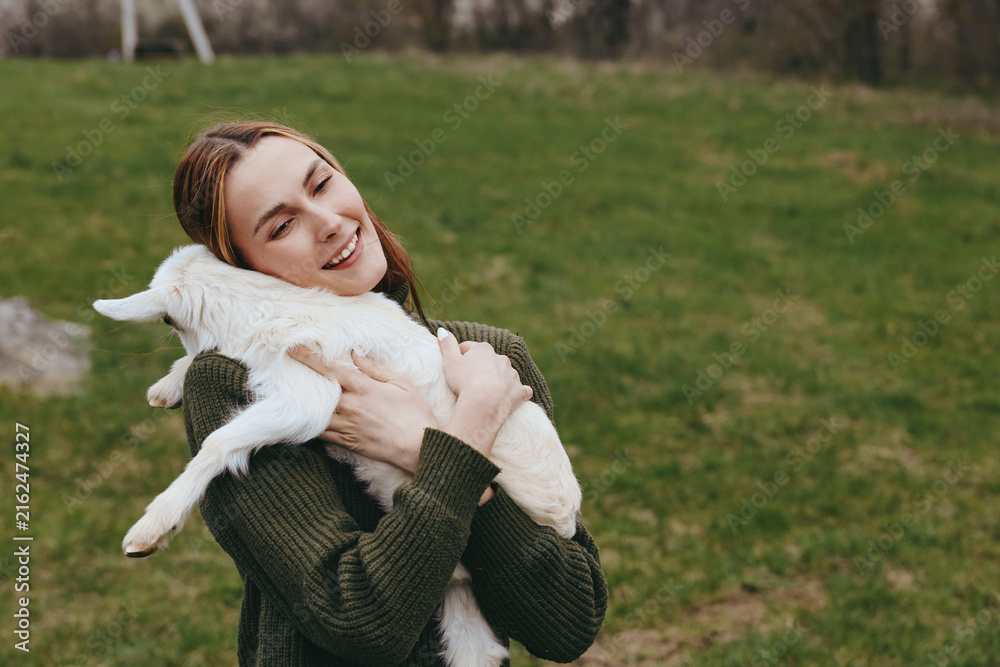 Fototapeta premium Joyful woman outdoors hugs a fluffy goat kid, sharing a tender moment in a grassy field, casual sweater, peaceful countryside mood, candid lifestyle scene, gentle interaction with animals.