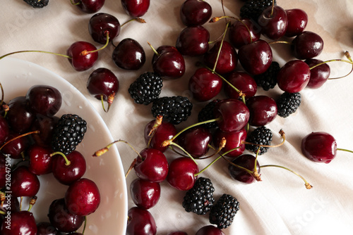 Cuadro en lienzo Fresh ripe sweet cherries and dark blackberries scattered on a white crumpled linen tablecloth with natural sunlight shadows